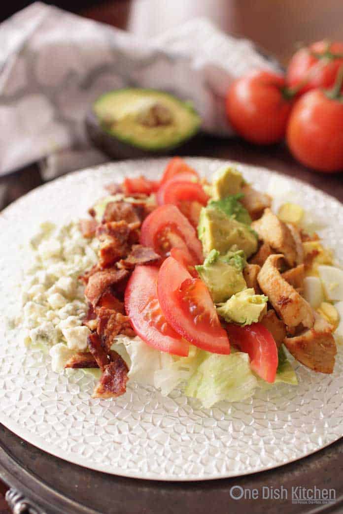 A cobb salad with blue cheese crumbles, crispy bacon bits, iceberg lettuce, tomato slices, avocado, hard boiled egg, and chicken on a large plate