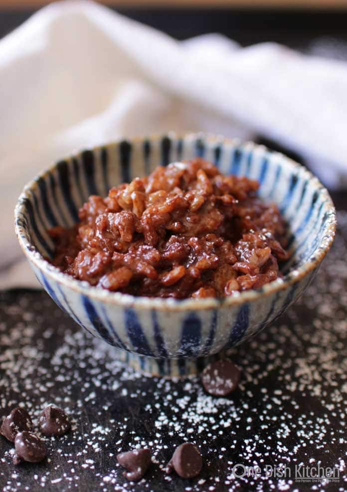 Chocolate rice pudding in a bowl on a dark surface with scattered chocolate chips and powdered sugar