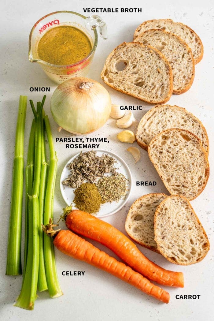 The ingredients for vegan stuffing: celery, carrots, bread, herbs, broth, garlic and onion, sitting on a white surface.