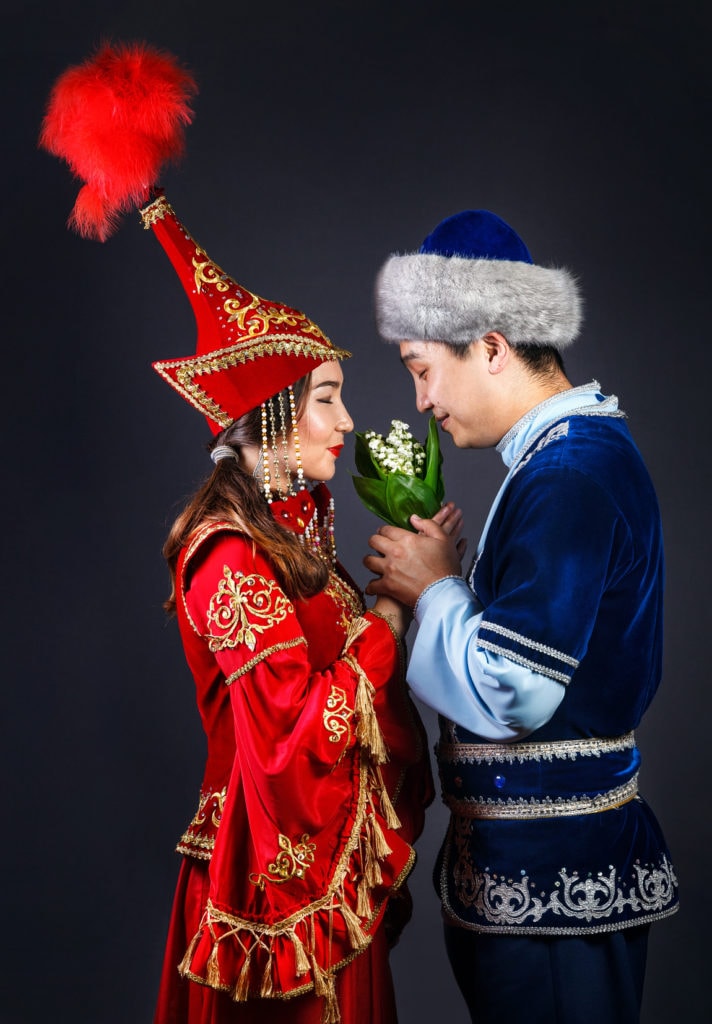Asian couple in national Kazakh costumes holding flowers and smell it at grey background