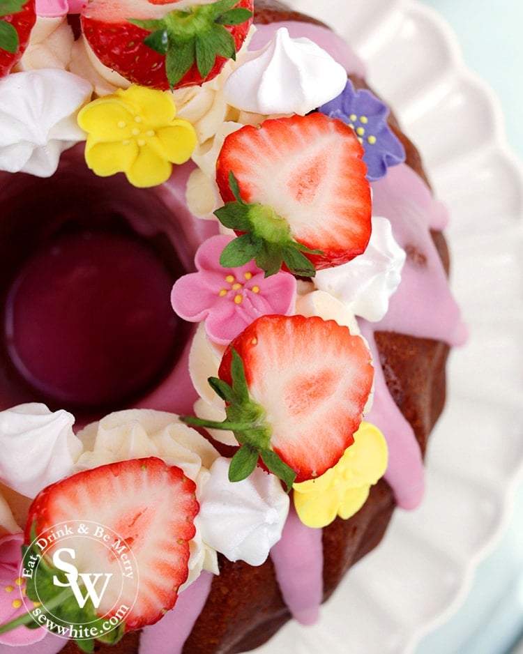 A close up of halved strawberries with mini meringue kisses and sugar flowers on bundt cake with pink icing.