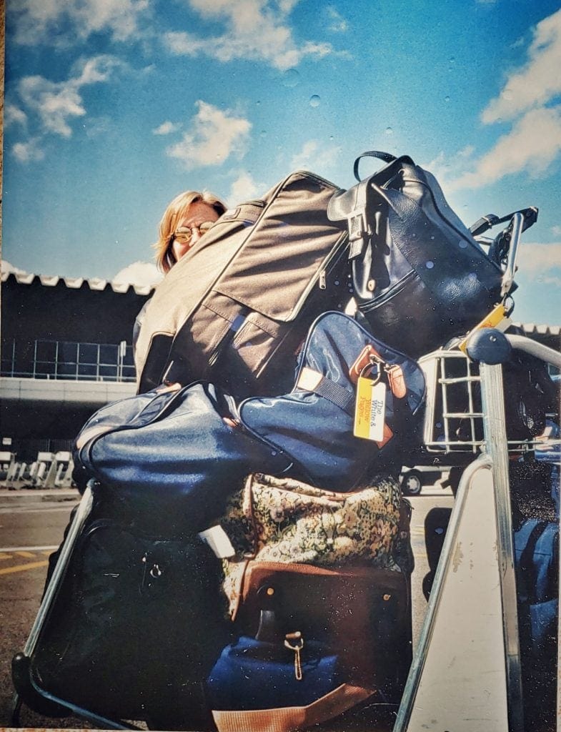 Vintage photo of author peeking from behind a huge pile of suitcases, handbags and wardrobe bags. She looks embarrassed at the Rome airport.