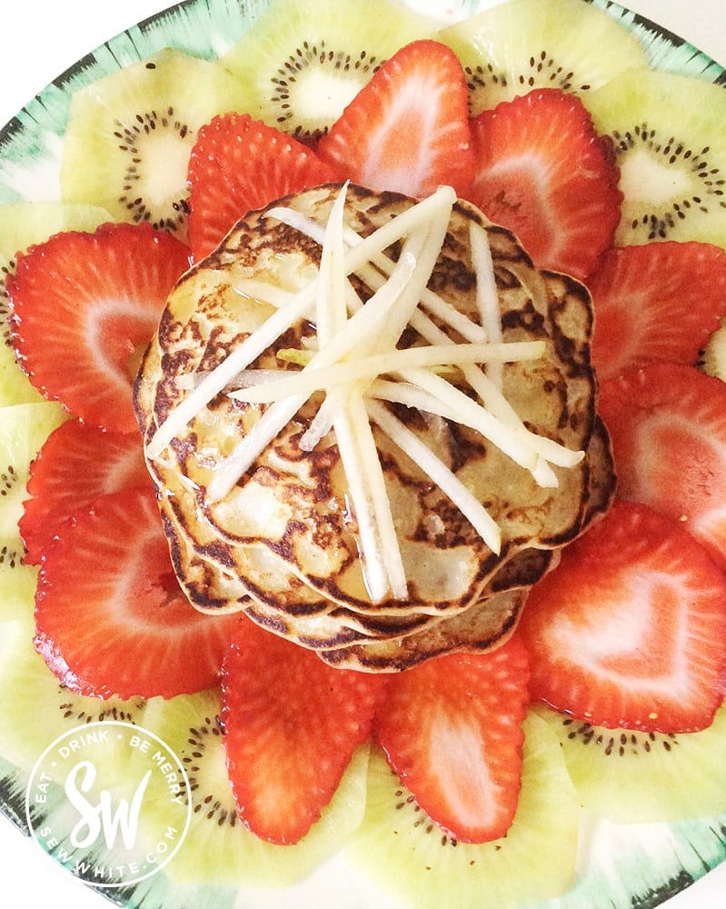 Apple pancakes topped with grated apple on top of a bed of fresh strawberries and kiwi.
