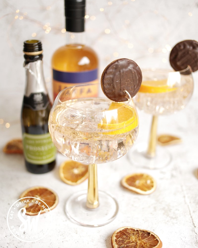 golden orange Jaffa Cake Gin Spritz in a gin glass decorated with jaffa cakes and surrounded by dried oranges.