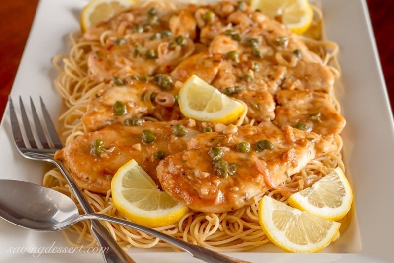 Try this classic Chicken Piccata Recipe made with tender chicken cutlets quickly sautéed then simmered in a light lemony, white wine sauce. Serve over pasta with capers and lemon wedges for an easy weeknight meal. #savingroomfordessert #chicken #chickenpiccata #easypiccata #chickenpiccatarecipe #chickencutlets #weeknightdinner #italian A platter of easy Chicken Piccata Recipe served with lemon wedges over hot spaghetti