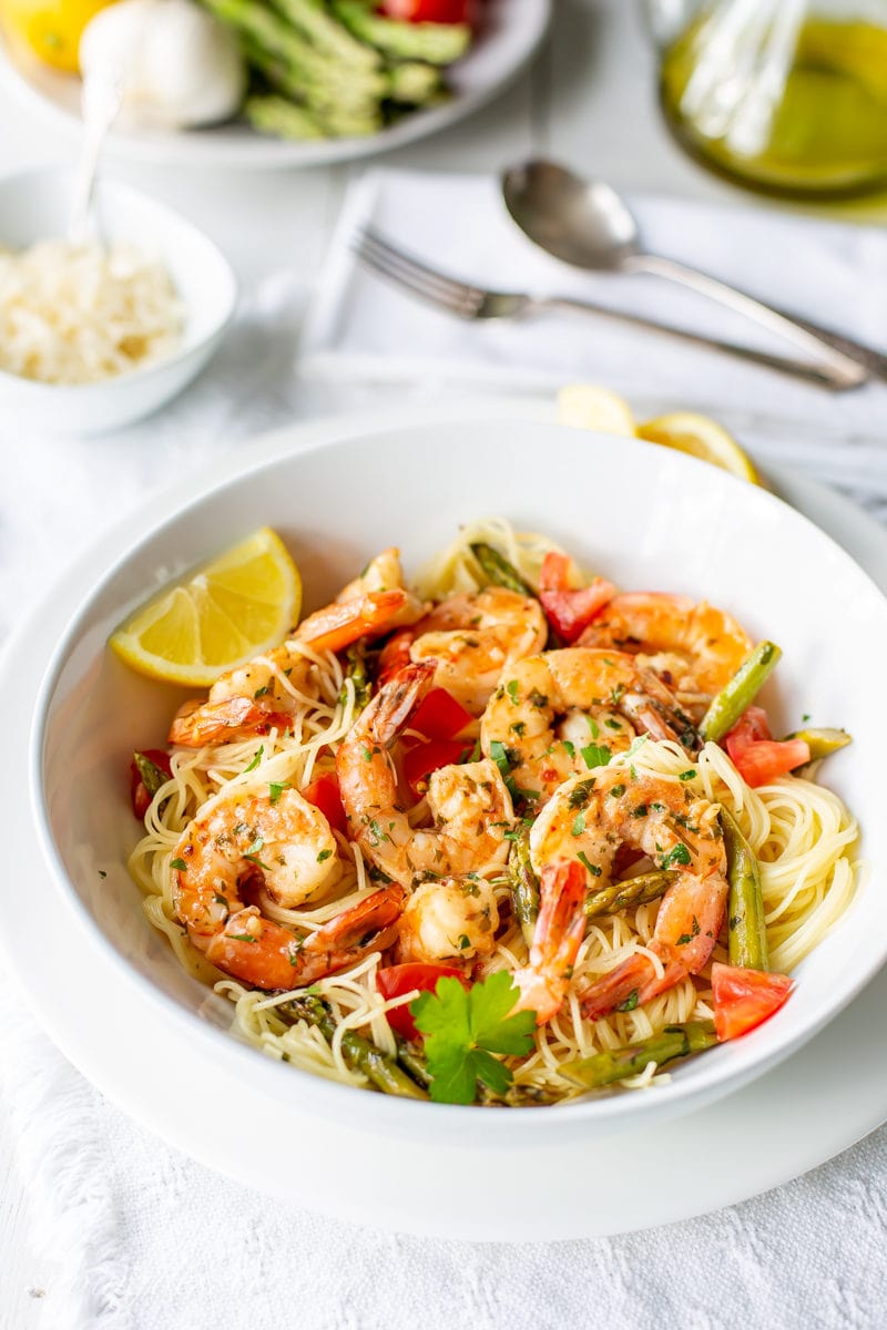 a bowl of shrimp scampi with lemon wedges, asparagus and tomatoes served over angel hair pasta