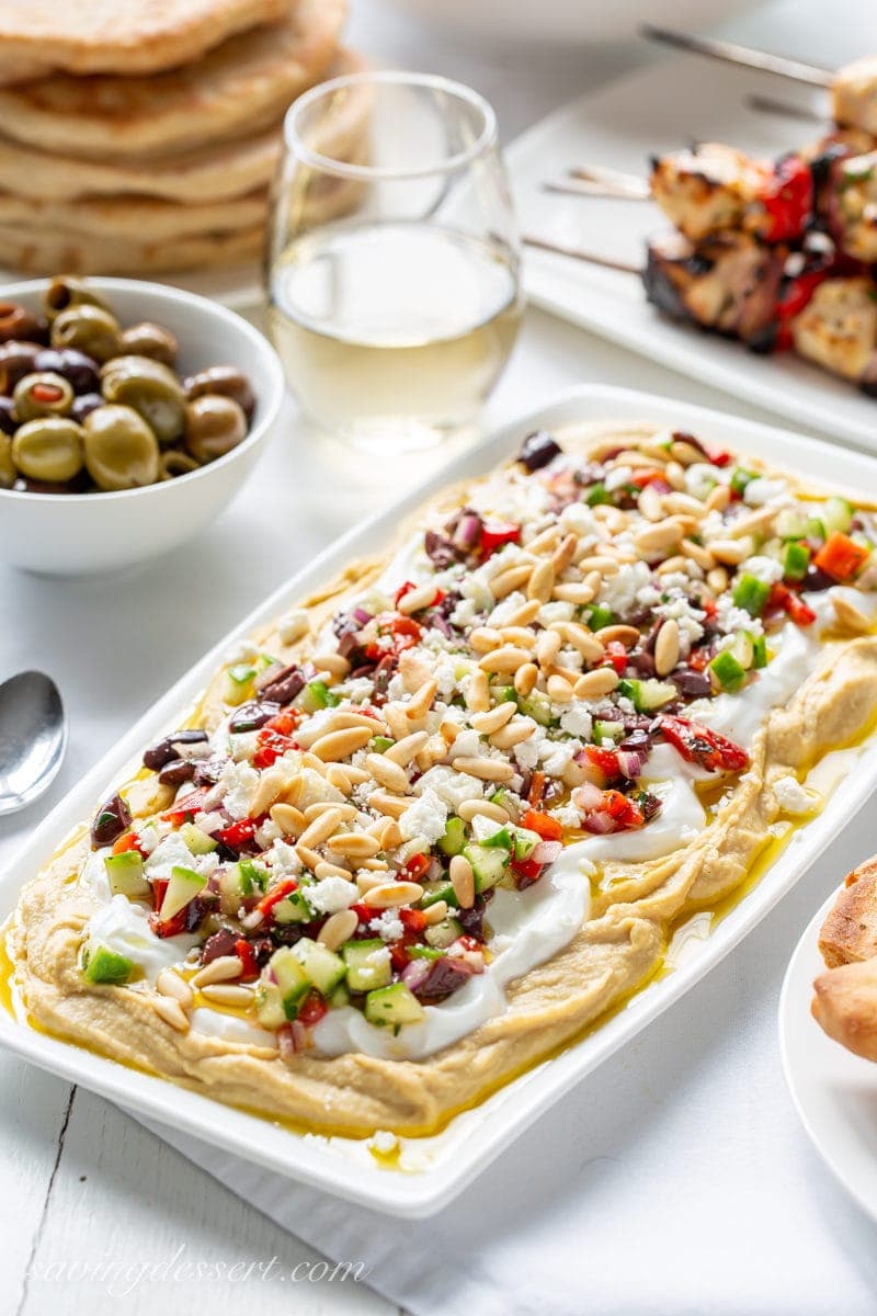 Enjoy this easy to make Greek inspired Layered Hummus Dip with tons of flavor and a great combination of textures; perfect for entertaining! #hummus #hummusdip #greekdip #layereddip #greeklayereddip #greekhummusdip #easyentertaining #greek A platter of Greek Layered Hummus Dip with yogurt, olives, red peppers, pine nuts and cucumbers