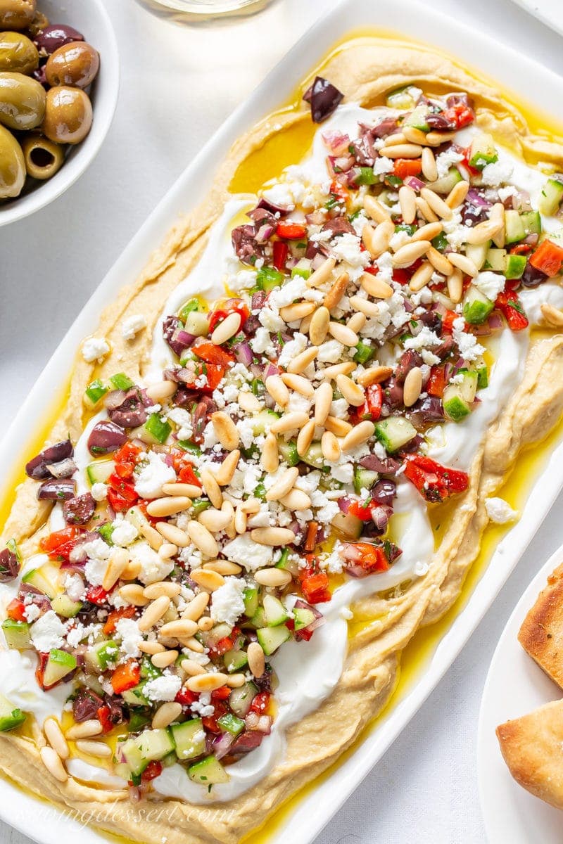 Enjoy this easy to make Greek inspired Layered Hummus Dip with tons of flavor and a great combination of textures; perfect for entertaining! #hummus #hummusdip #greekdip #layereddip #greeklayereddip #greekhummusdip #easyentertaining #greek A small platter with a Greek Layered Hummus Dip with yogurt, red peppers, cucumbers, onion, olives, feta cheese and pine nuts