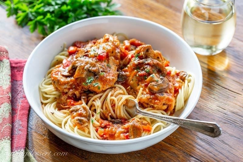 Really good Chicken Cacciatore is one of the most satisfying, delicious and comforting Italian dishes you can make at home. Great served with spaghetti or over polenta. #savingroomfordessert #chicken #chickenthighs #chickencacciatore #cacciatore #comfortfood #italiandish #italian A bowl of spaghetti topped with Chicken Cacciatore with mushrooms, red peppers and parsley served with a glass of white wine