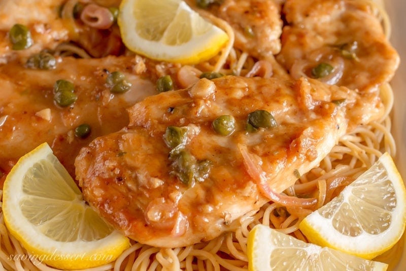 Try this classic Chicken Piccata Recipe made with tender chicken cutlets quickly sautéed then simmered in a light lemony, white wine sauce. Serve over pasta with capers and lemon wedges for an easy weeknight meal. #savingroomfordessert #chicken #chickenpiccata #easypiccata #chickenpiccatarecipe #chickencutlets #weeknightdinner #italian A closeup of chicken piccata with capers and lemon wedges served over spaghetti
