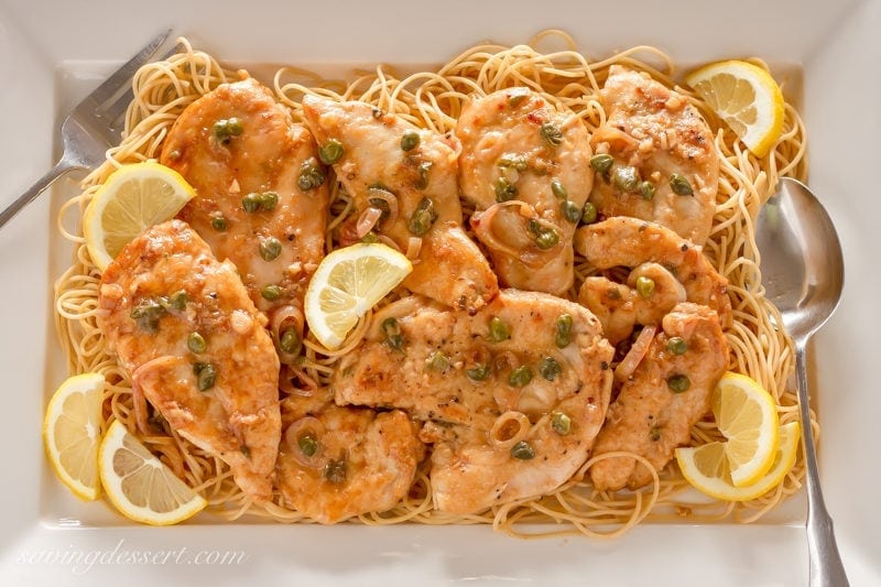 Try this classic Chicken Piccata Recipe made with tender chicken cutlets quickly sautéed then simmered in a light lemony, white wine sauce. Serve over pasta with capers and lemon wedges for an easy weeknight meal. #savingroomfordessert #chicken #chickenpiccata #easypiccata #chickenpiccatarecipe #chickencutlets #weeknightdinner #italian A platter of Chicken Piccata over spaghetti garnished with lemon wedges