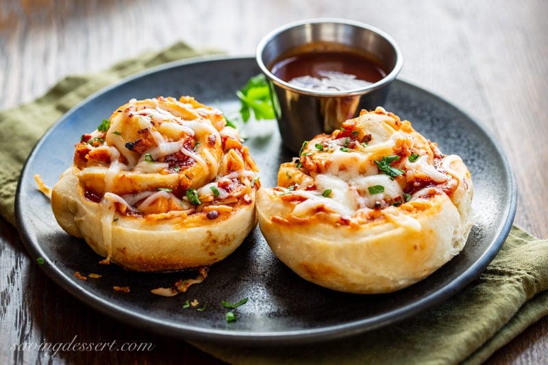 Bring on the party! I've got the BEST recipe for amazing Cheesy BBQ Chicken Pizza Rolls right here #savingroomfordessert #pizza #pizzarolls #bbqchickenpizza #bbqpizza #appetizers #partyfood A plate with two Cheesy BBQ Chicken Pizza Rolls with a cup of BBQ sauce on the side