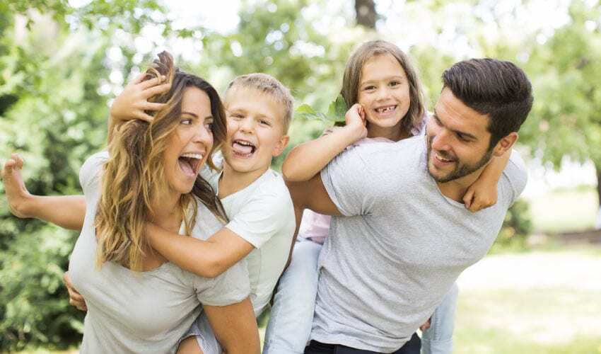 Wife and husband give piggy-back rides to their son and daughter while smiling and laughing outside