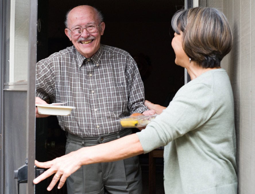 Meals on Wheels People representative delivers food to an elderly man at his front door.