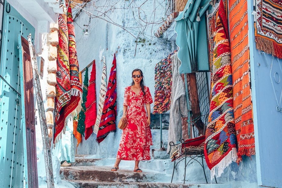 What Is The Cost Of Travelling In Morocco 18 L Jasmine of The Travel Quandary poses in front of woven Moroccan rugs for sale, Chefchaouen Morocco