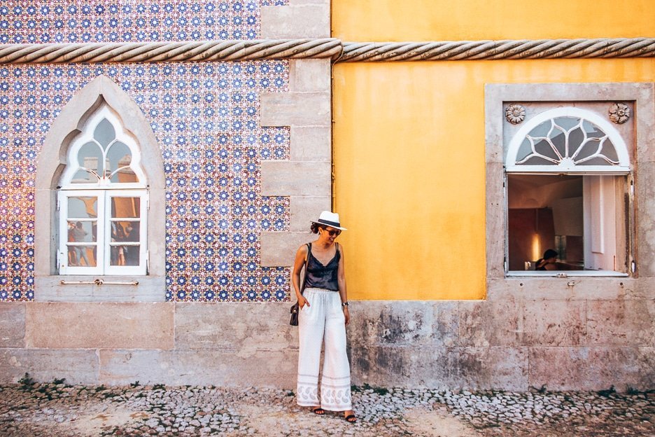 About Us 2 L A woman poses in front of the yellow and blue walls of Pena Palace in Sintra, Portugal