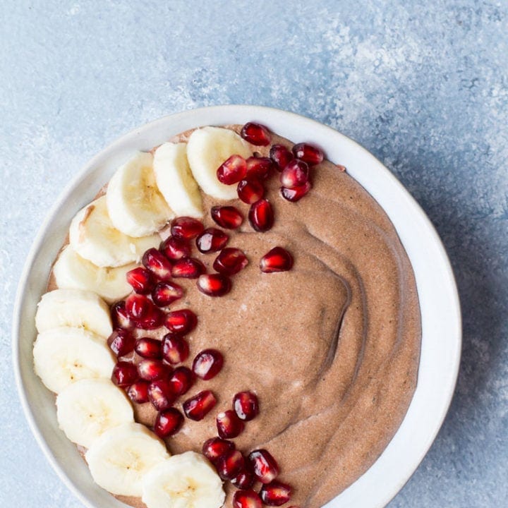 Bowl with chocolate chia pudding, decorated with banana slices and pomegranate to the left. Blue background, flatlay.
