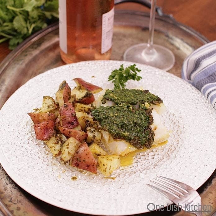Baked Cod topped with chermoula with roasted chopped red potatoes on a glass plate and a bottle of rosé in the background all on a metal tray