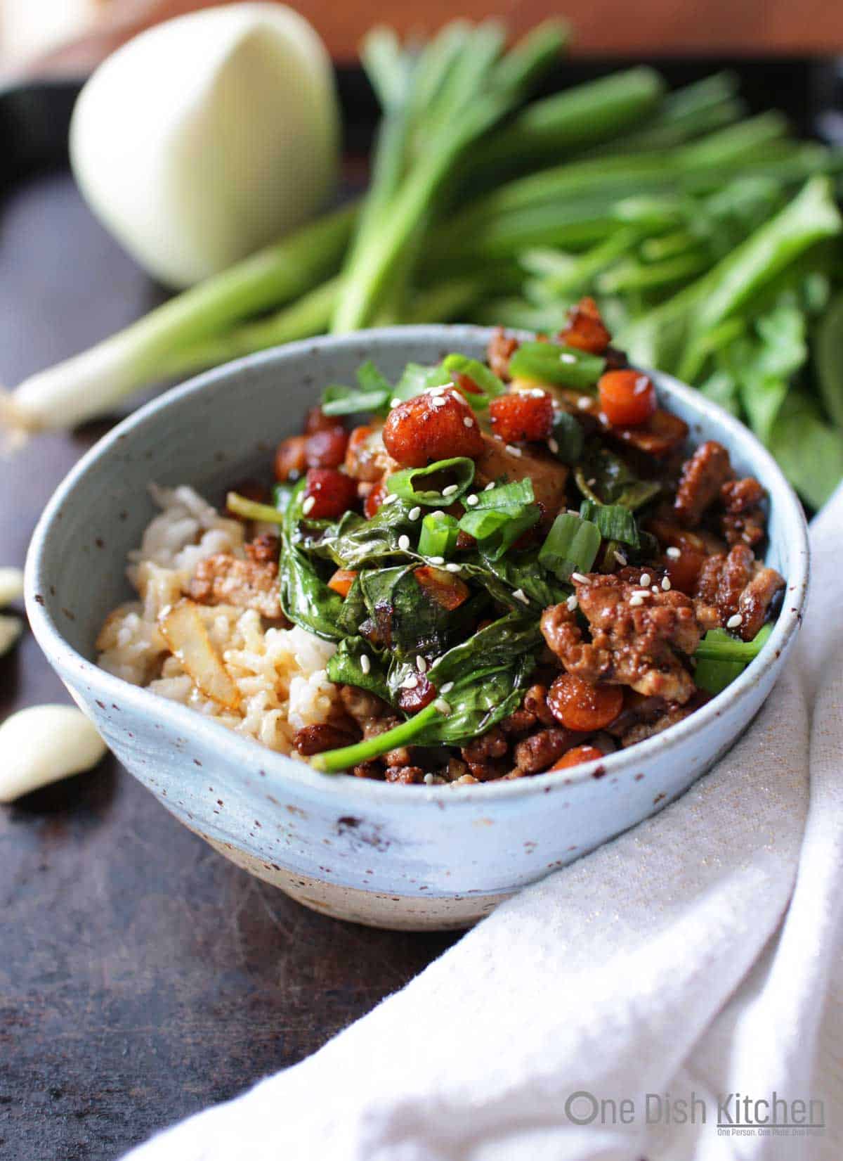 A bowl of pork stir fry over white rice surrounded by a white cloth napkin, fresh green onions, spinach, and garlic cloves all on a metal tray