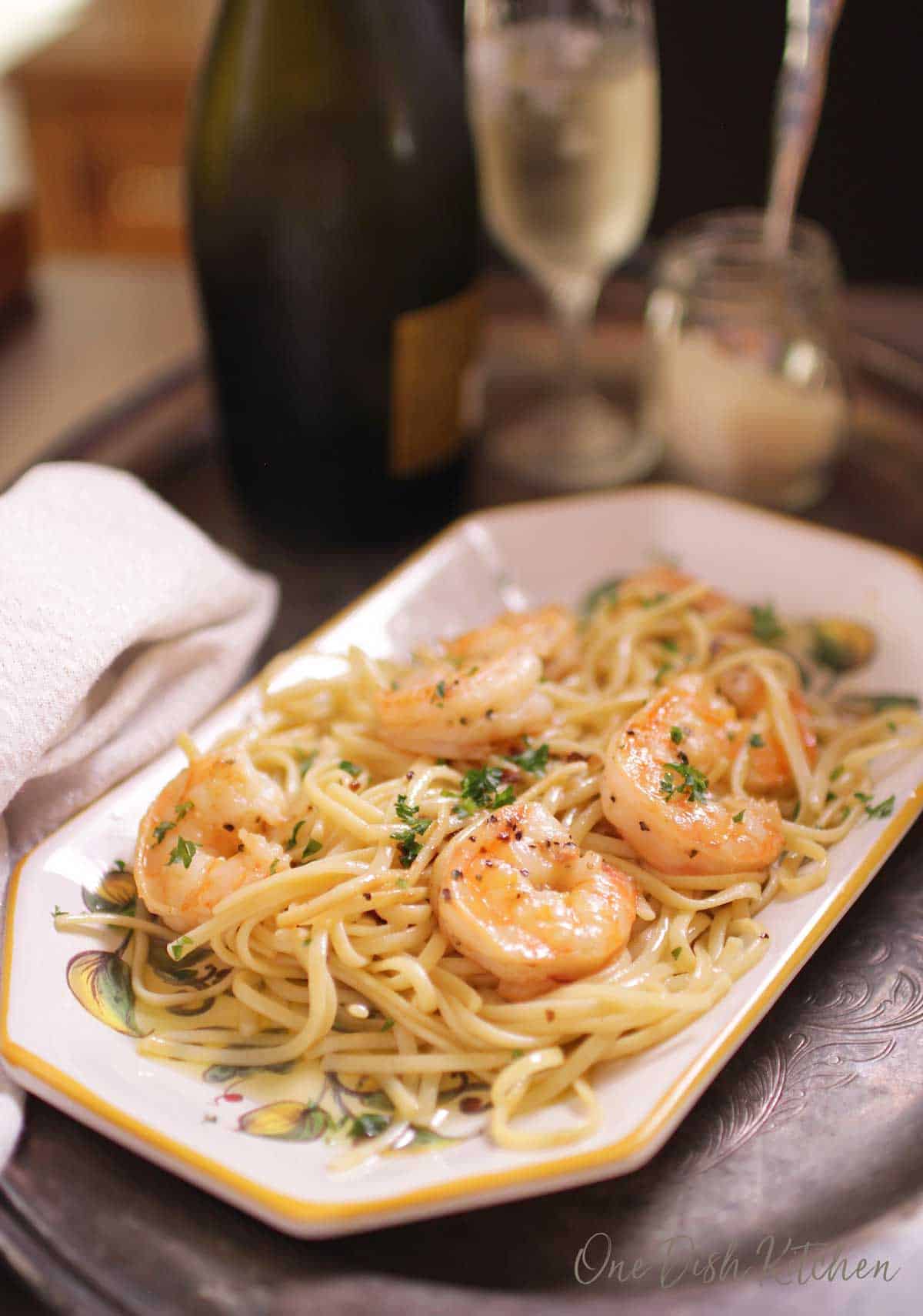A plate of spaghetti with shrimp on a tray next to a jar of parmesan cheese and a glass of champagne.