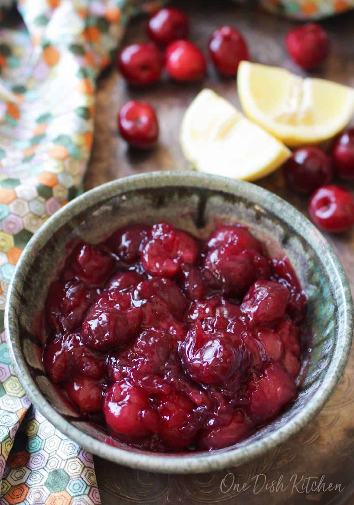 An overhead view of a bowl of cherry pie filling next to a colorful napkin and scattered cherries and lemon slices all on a metal tray
