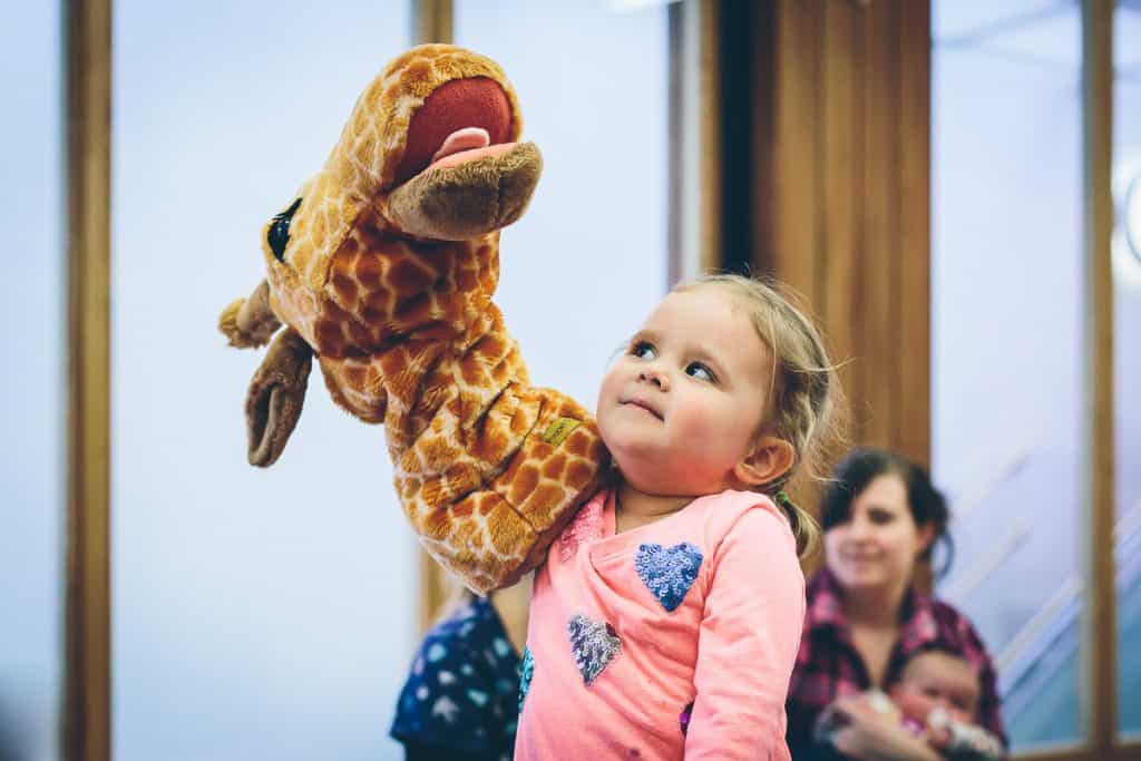 A young girl with her arm inside a giraffe hand puppet.