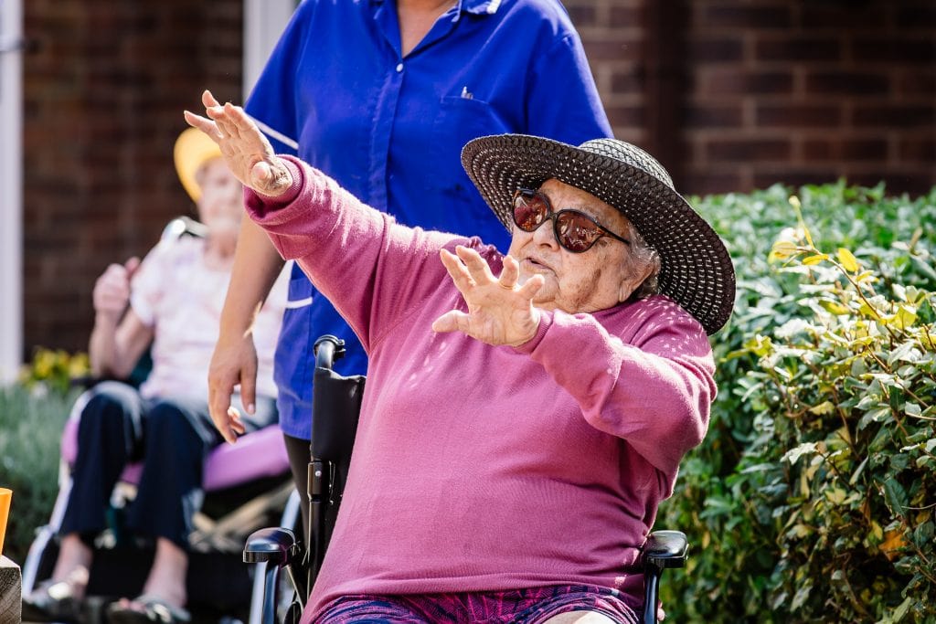 A woman in a hat and glasses dances with her arms in the air while sitting in a wheelchair at a dance session led by darts in Doncaster