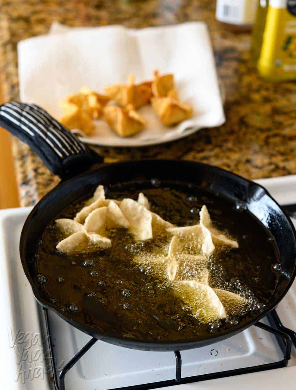 Frying vegan Crab Rangoon in a large, cast iron skillet, on a the stove, with cooked pieces on a plate in the background.