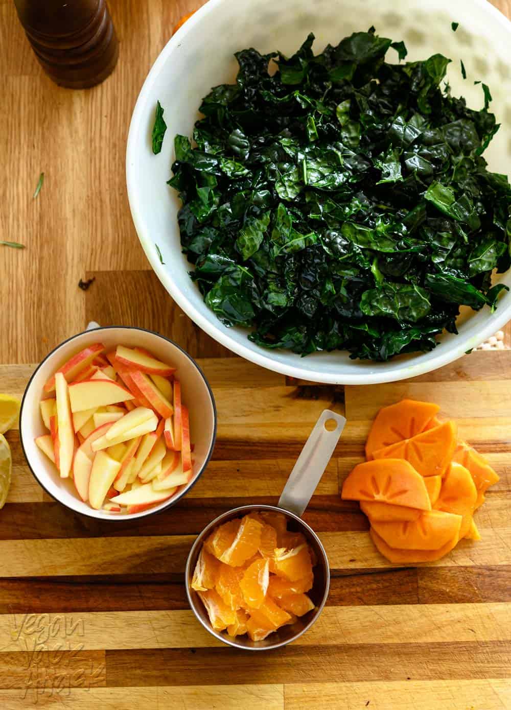 Bowls filled with kale, apples, tangerine pieces, and persimmon slices, on a cutting board