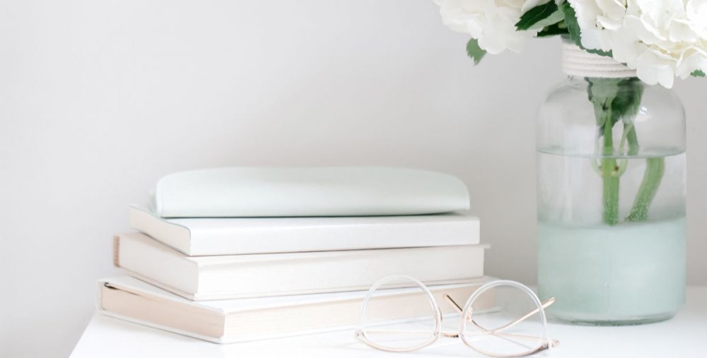stack of books on nightstand with reading glasses and vase of white flowers