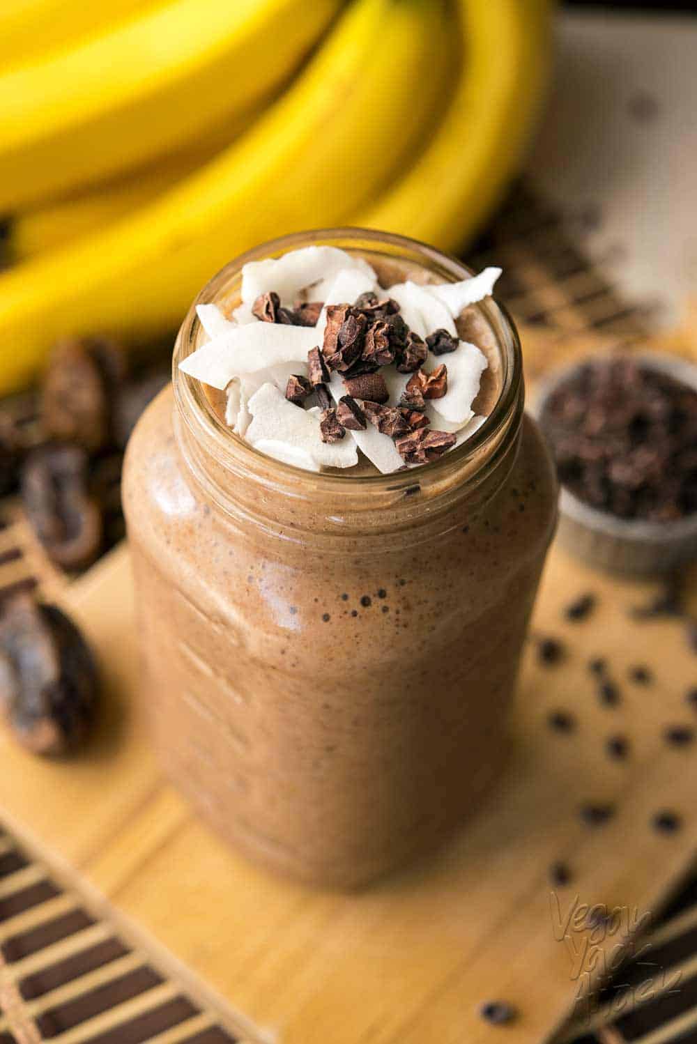 Large jar filled with banana cacao recovery smoothie, on a bread board surrounded by ingredients