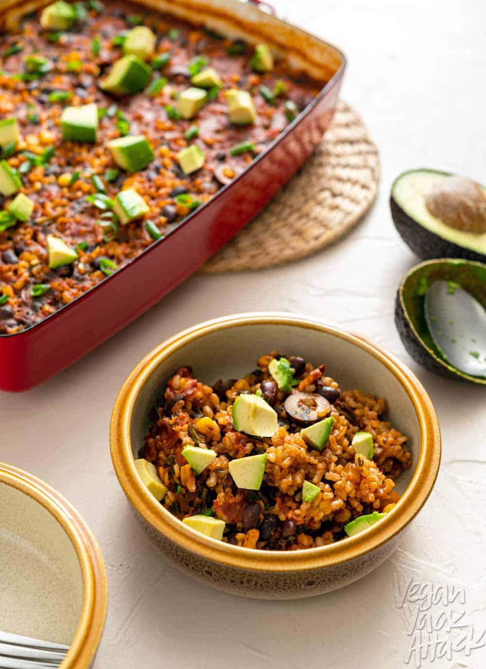 Image of chipotle brown rice bake in a red casserole dish on a beige background with bowl in foreground