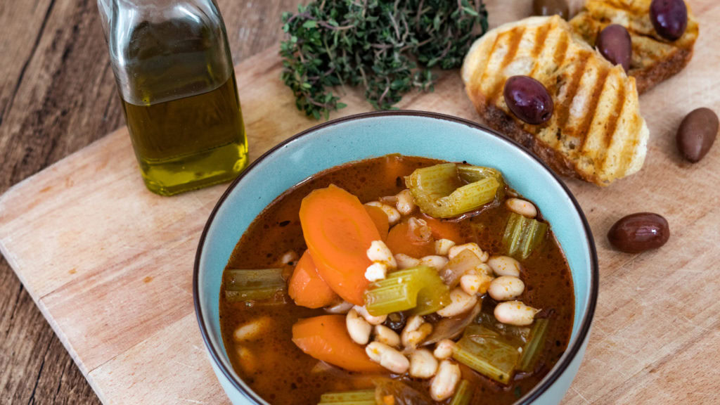 Buntes Gemüse in einer roten Bohnensuppe auf einem Holzbrett. Brot, Oliven, Thymian und Olivenöl im Hintergrund.