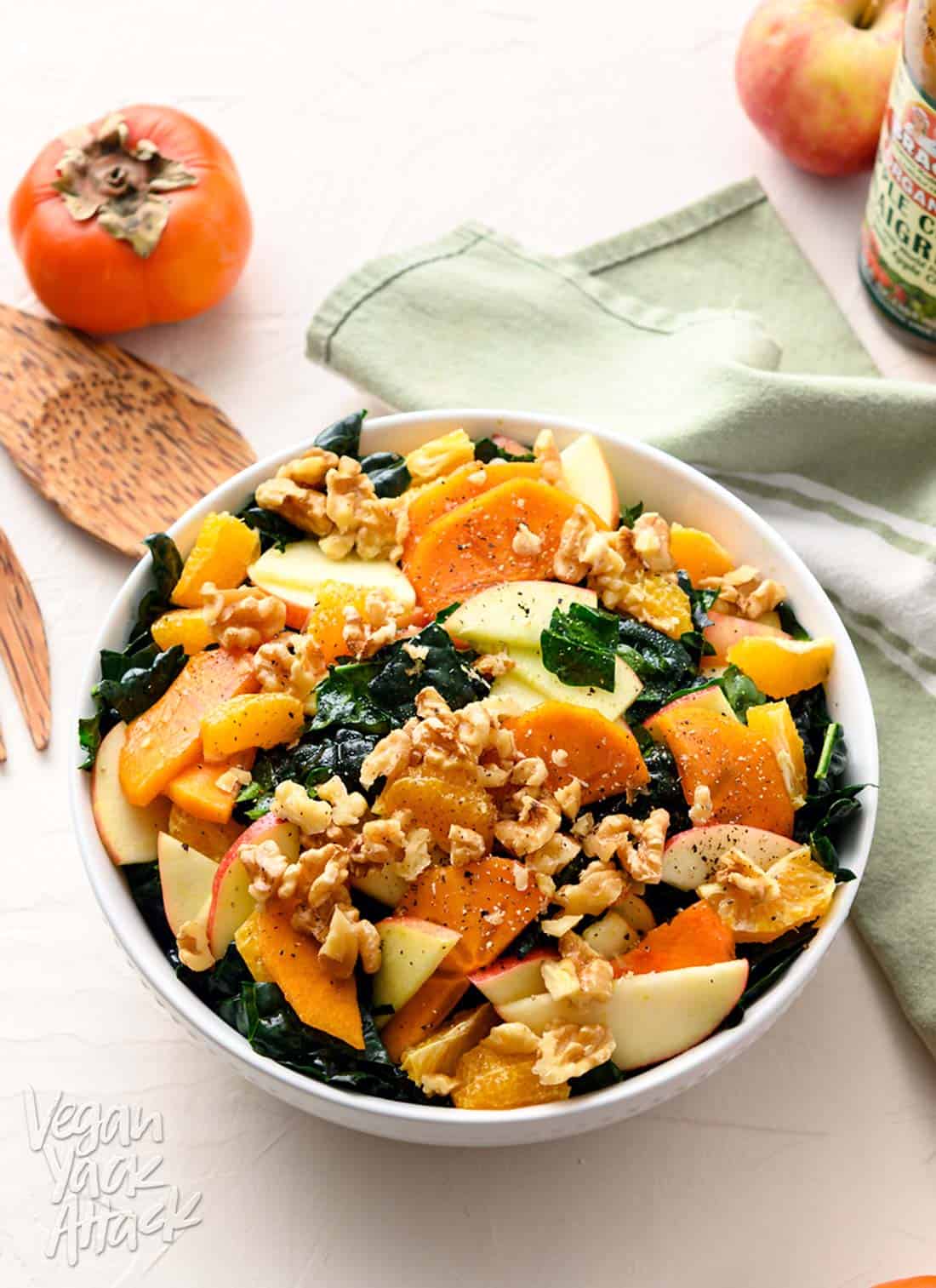 Persimmon apple kale salad in a large white bowl with cloth, salad utensils, and fruits next to it.