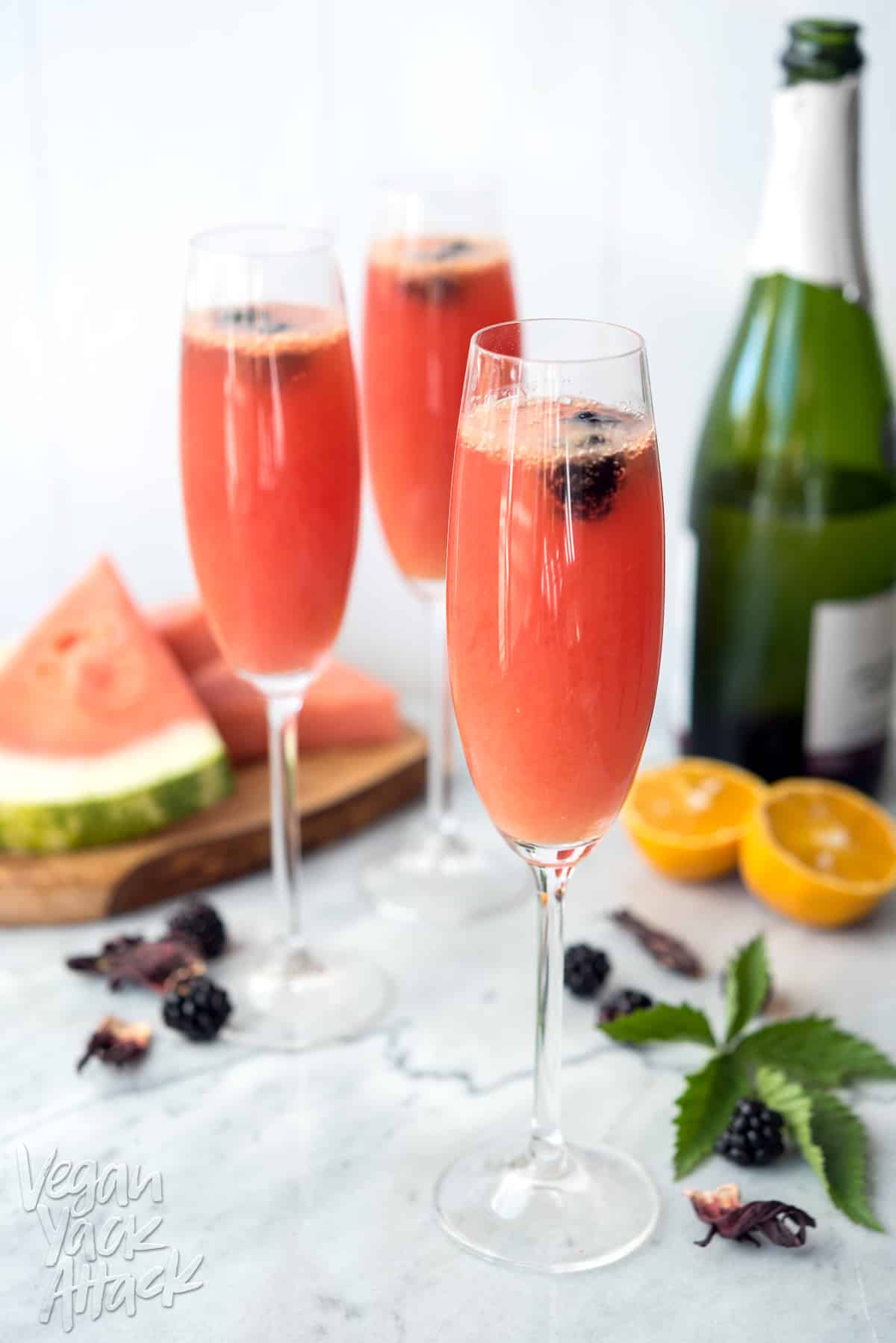 Image of three Champagne glasses filled with watermelon hibiscus mimosas, on a marble table top, surrounded by ingredients.