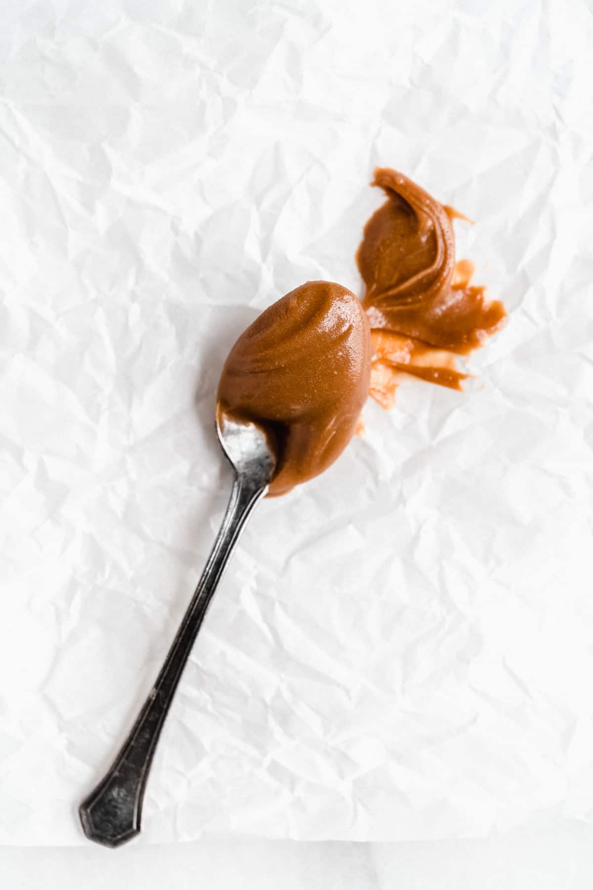 Overhead closeup photo of a silver spoon full of the 3-Ingredient Vegan Caramel Sauce. Spoon is sitting on white parchment paper and some of the caramel sauce has been spread onto the paper.