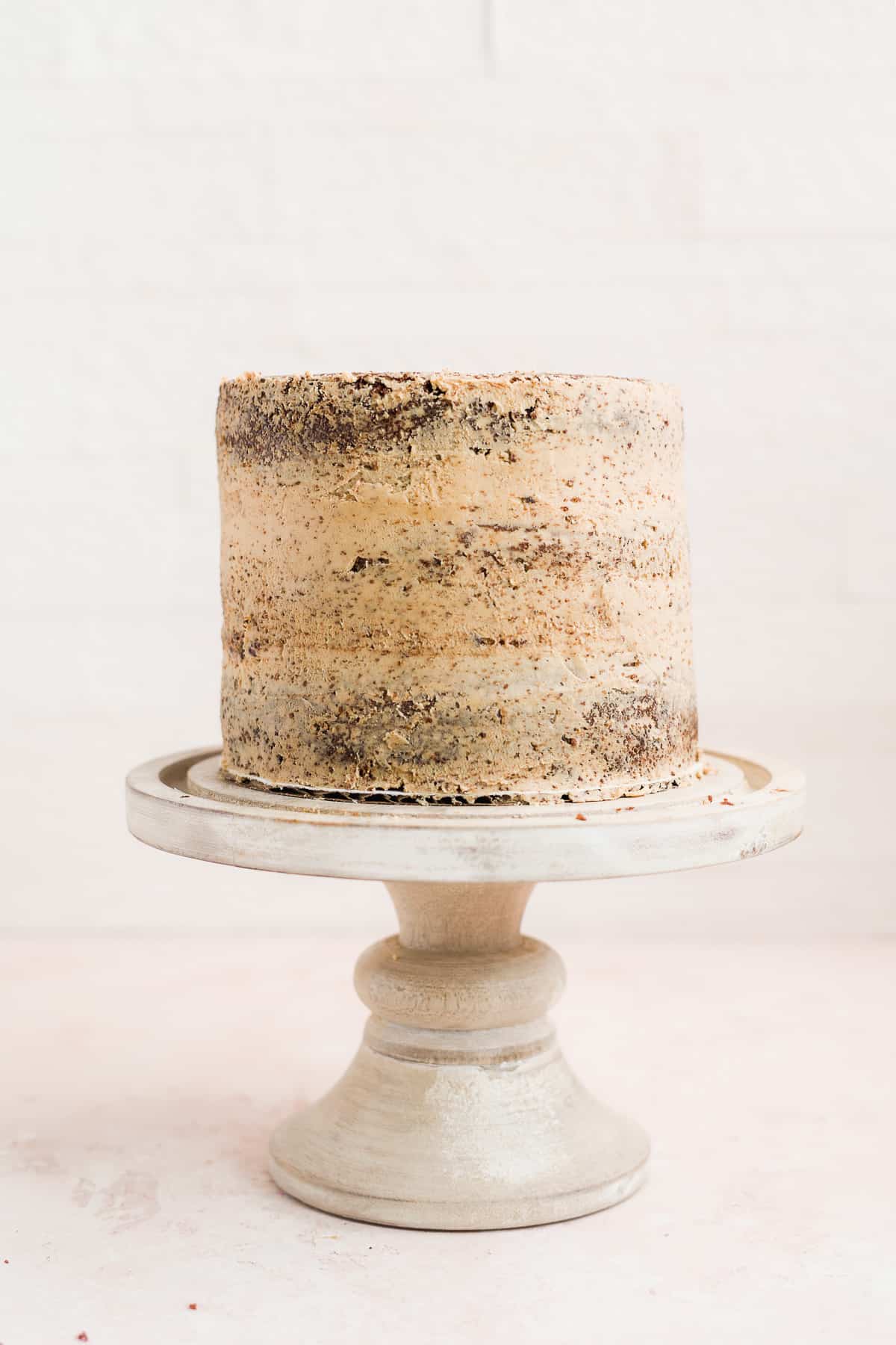 Cake on a stand with crumb coat on a pink surface.