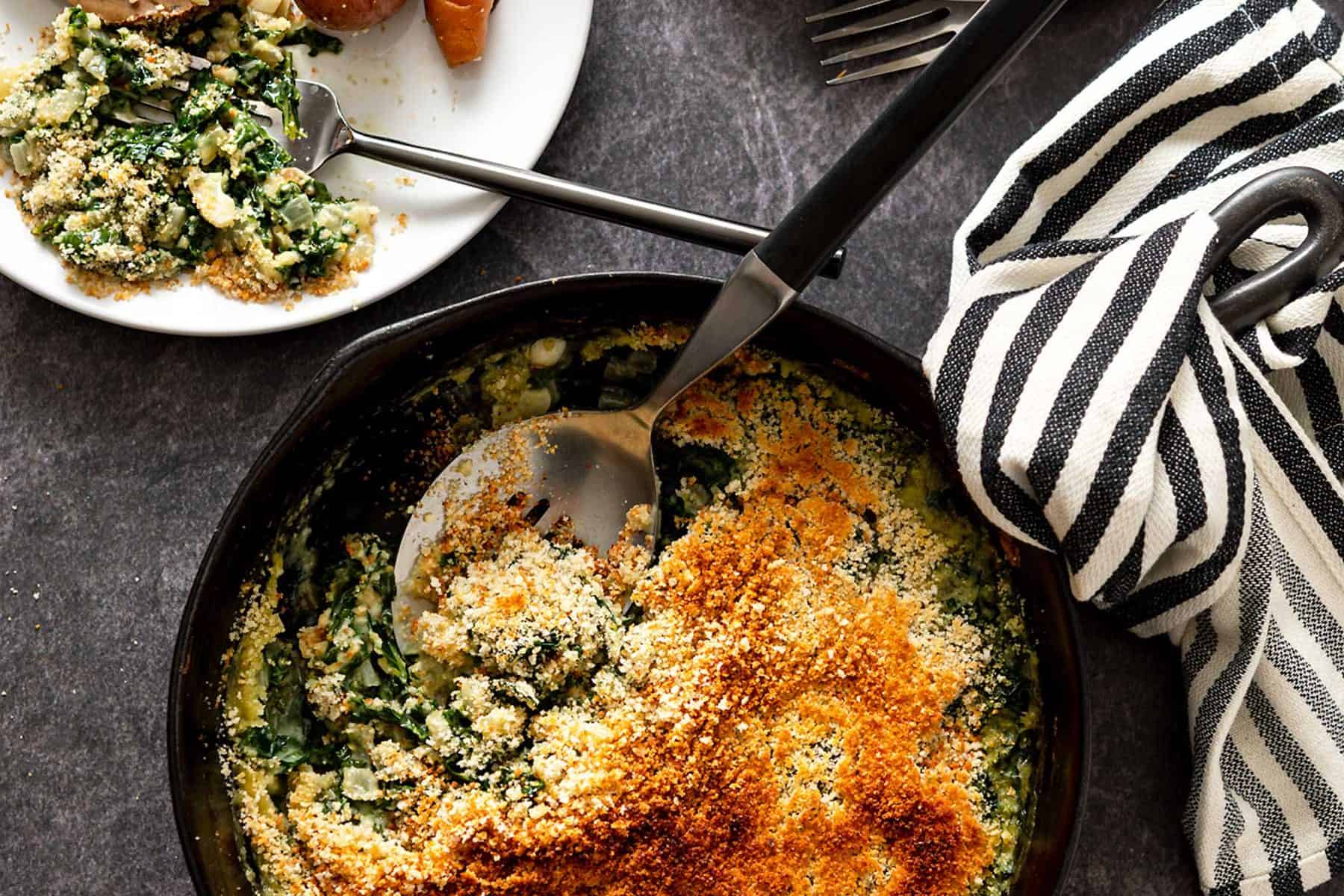 Serving spoon in a cast iron skillet filled with creamed kale next to dinner plates