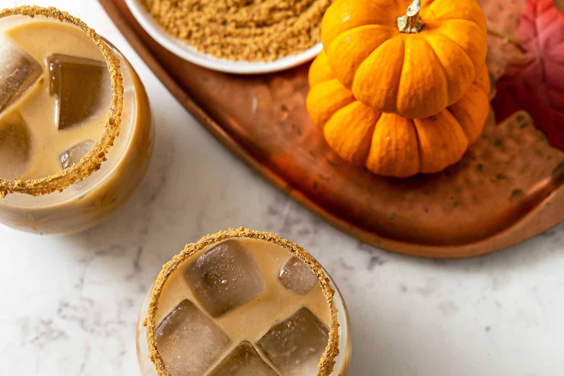 Two glasses filled with a tan-colored cocktail and pumpkins and crushed graham crackers around them