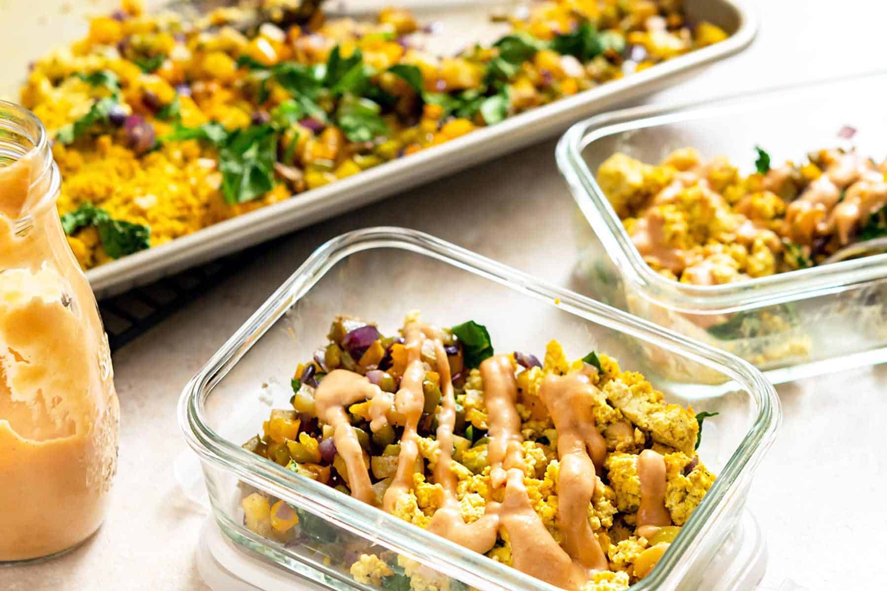 Two meal prep containers filled with Sheet Pan Tofu Scramble and hash, drizzled with sauce, next to a sheet pan