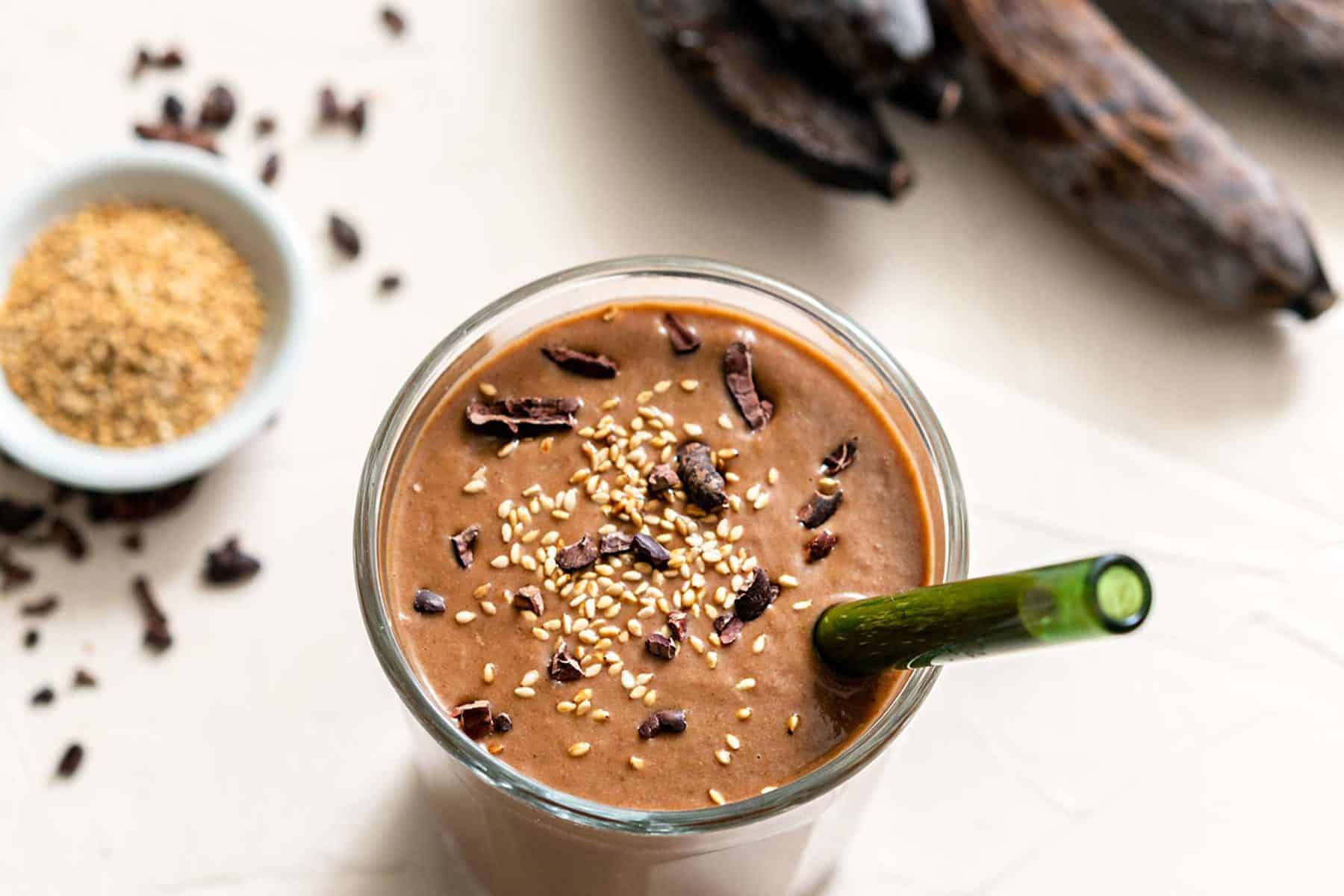 One large glass filled with chocolate sesame smoothie, with seeds, nibs, and bananas on the side