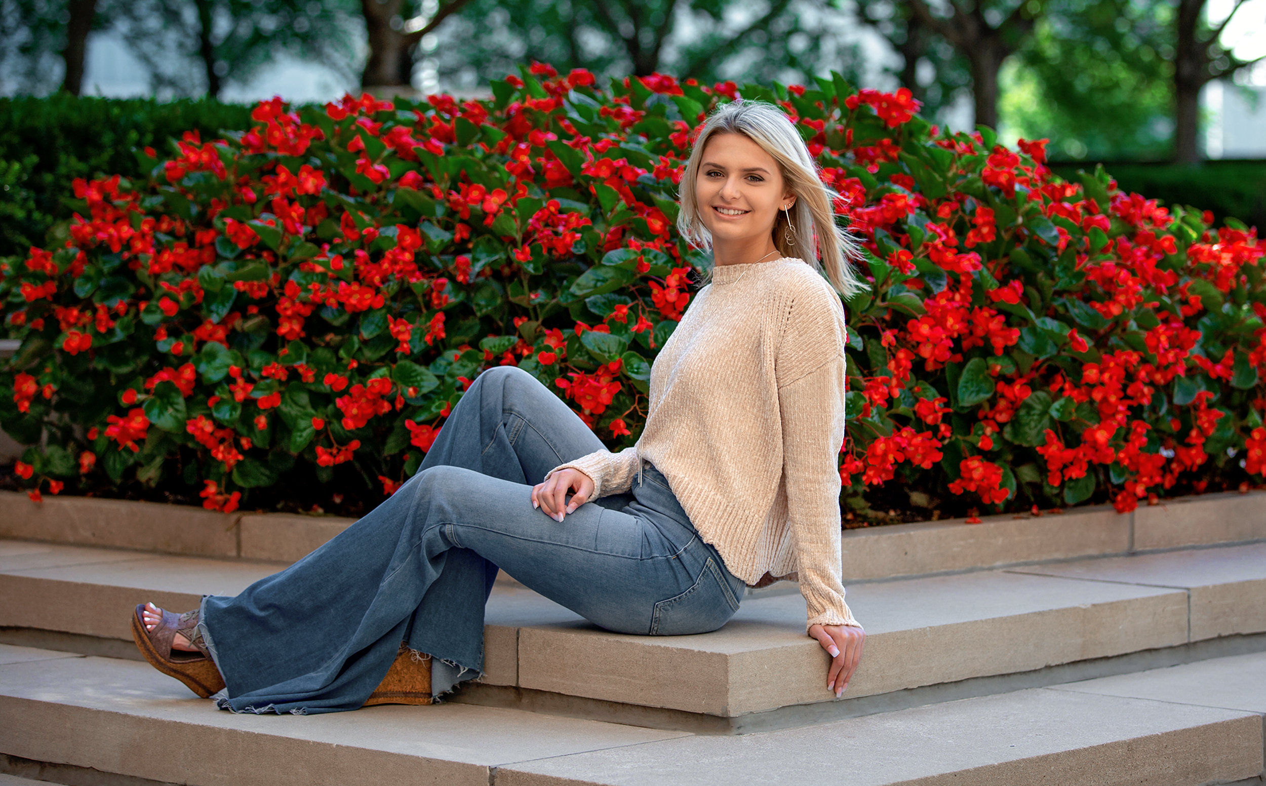Kidrowski_Creative_senior_photography_Nelson_Atkins_Museum A young blonde girl wearing bell-bottom jeans and a pink sweater sitting three-quarter profile to the camera on a small set of stairs with vibrant red flowers in the background.