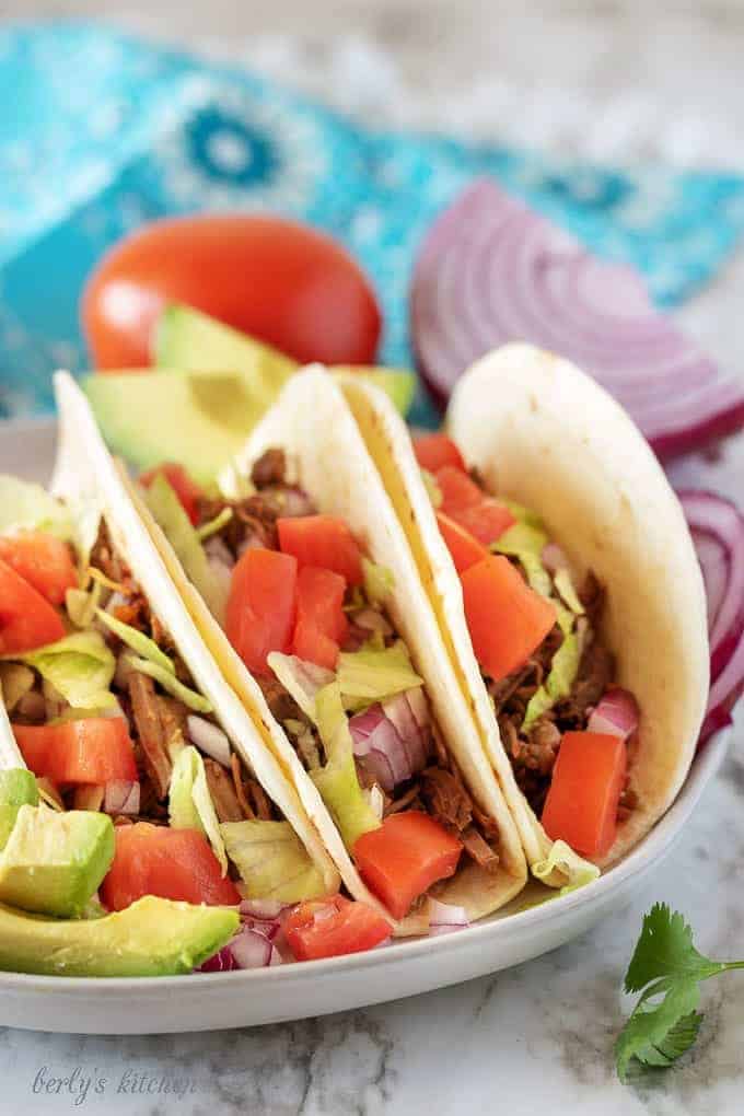 Three shredded beef tacos on a plate next to a tomato and purple onion.