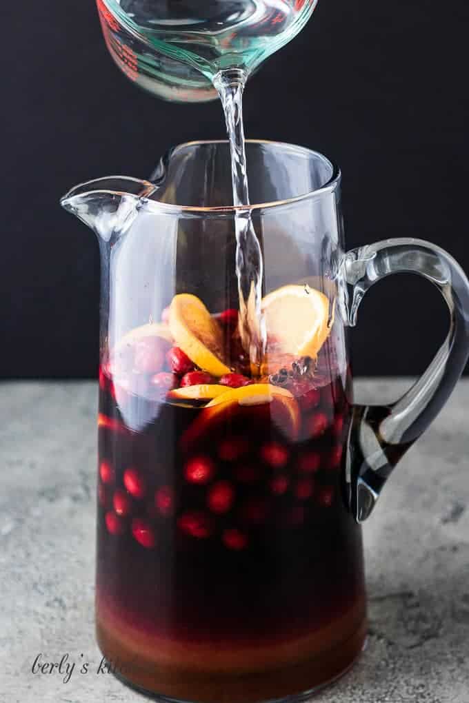 Club soda being poured into the pitcher filled with fruit and brandy.