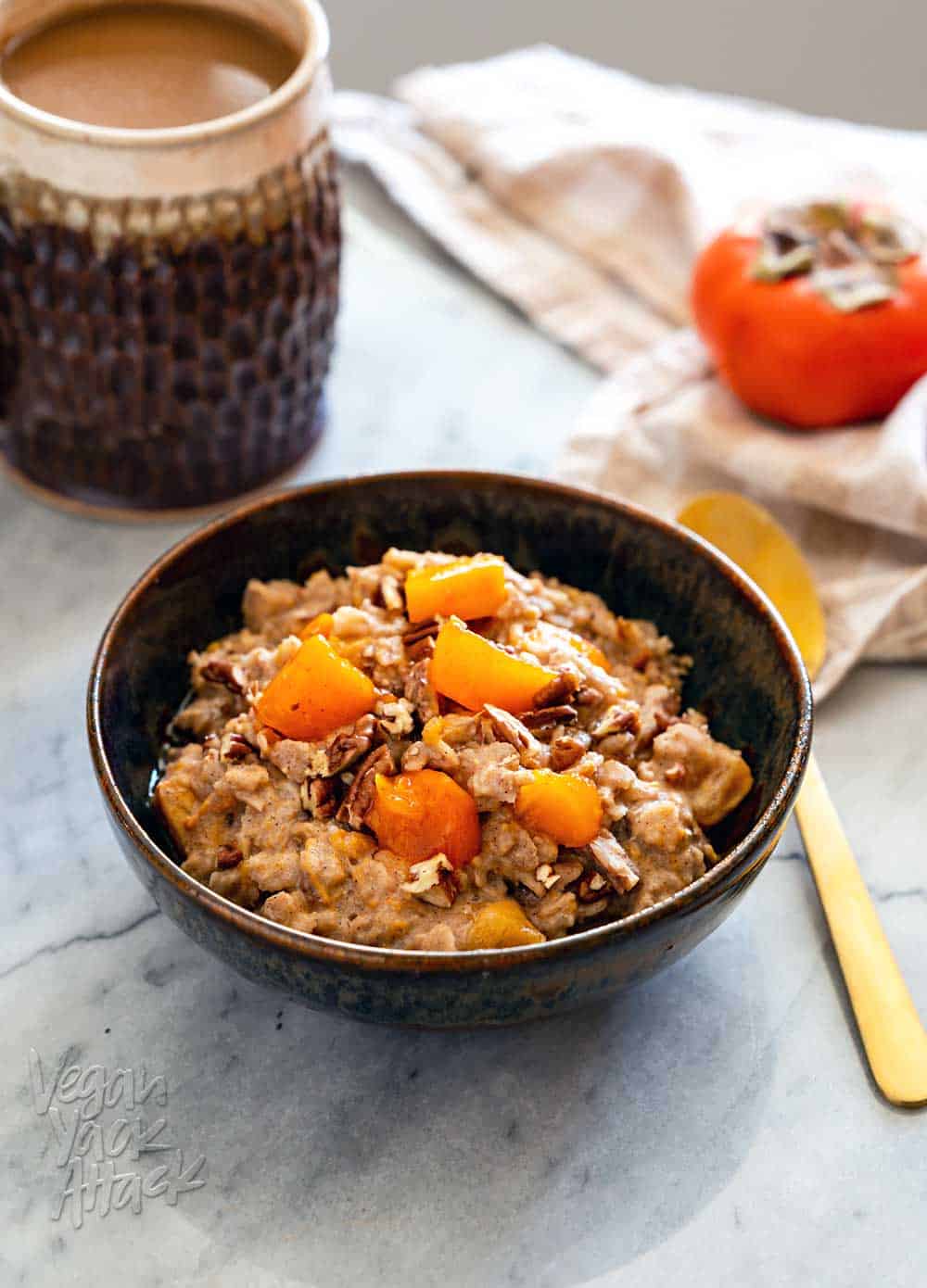 Pecan Persimmon Oatmeal Ceramic bowl filled with persimmon oatmeal, and golden spoon, with a cup of coffee