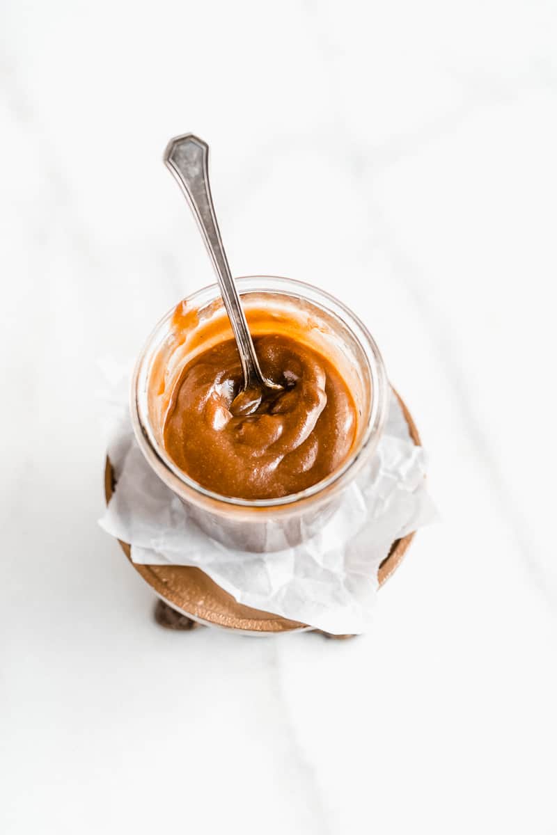 Overhead photo of a jar of the 3-Ingredient Vegan Caramel Sauce. A spoon is standing up inside the jar and the jar is sitting on white parchment paper.