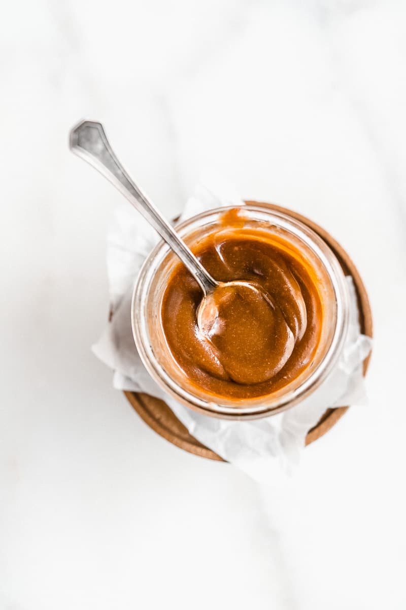 Overhead photo of a jar of the 3-Ingredient Vegan Caramel Sauce sitting on a small cake plate. A silver spoon is sticking up from the jar of caramel sauce.
