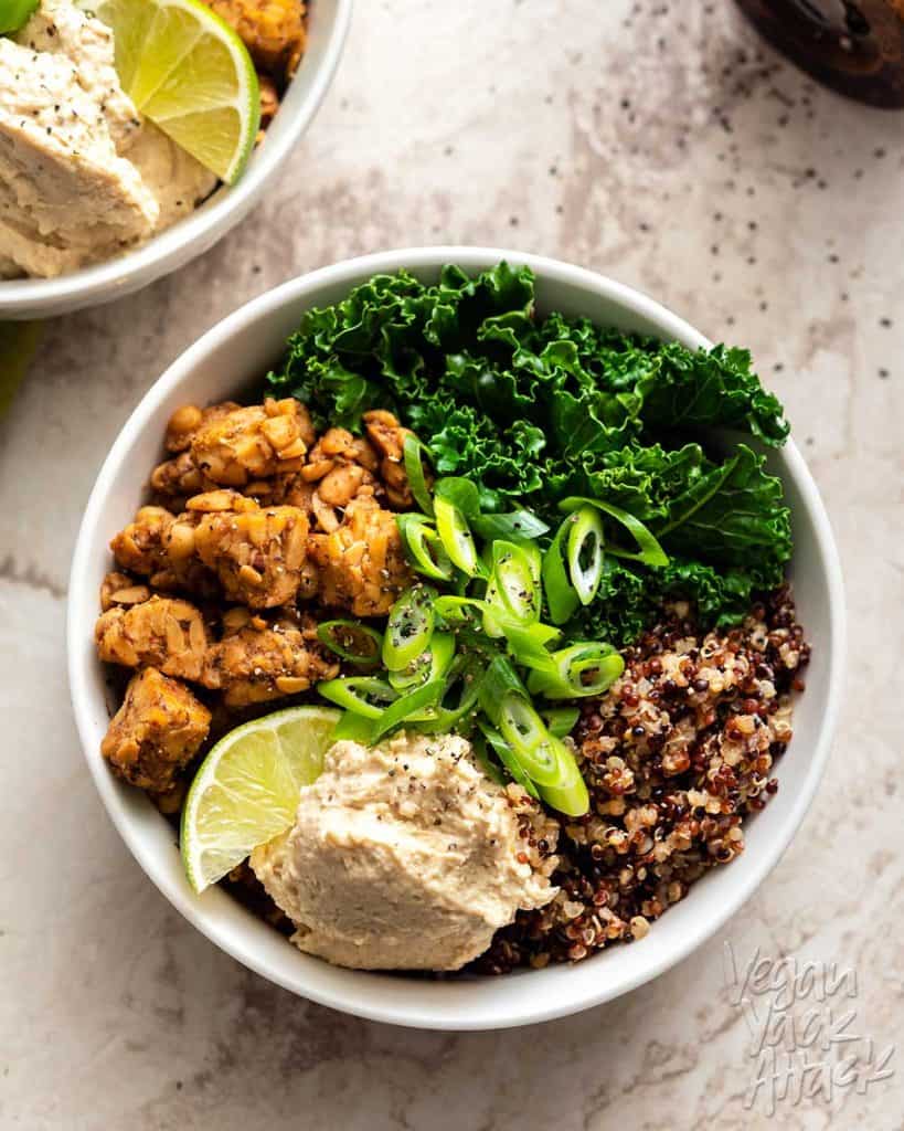 Two white bowls with kale, tempeh, quinoa, and bean dip on a marble background