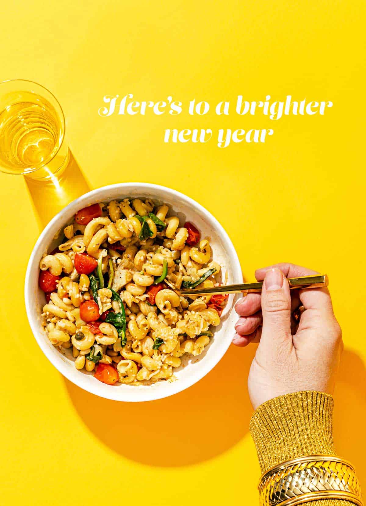 Bowl of pasta with hand holding fork, and champagne flute on a yellow background