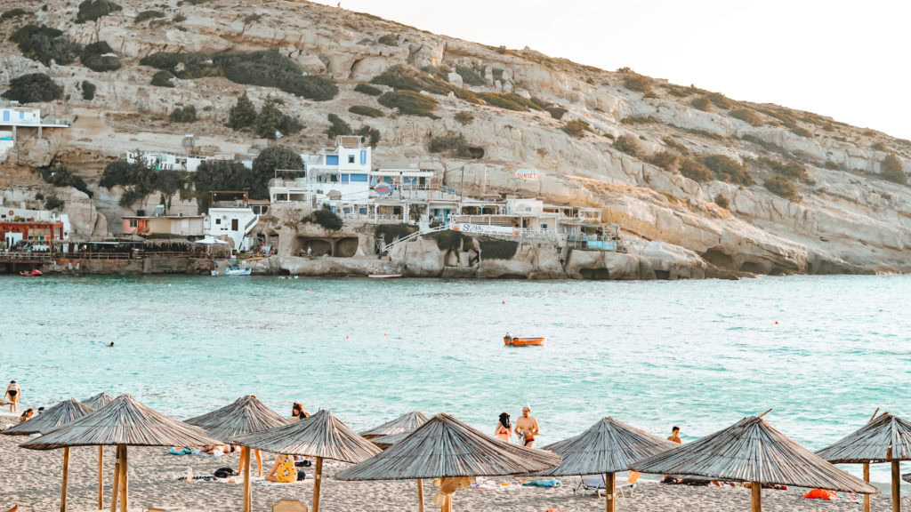 Sandstrand in Matala mit Schirmen und kleinen Häusern im Hintergrund